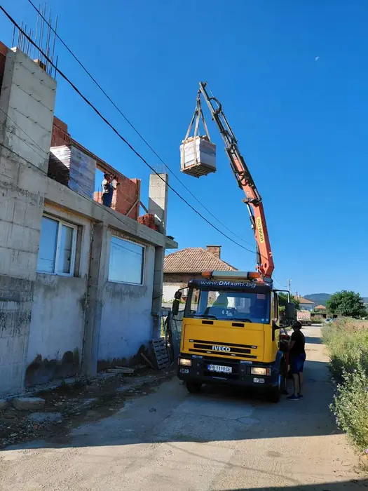 Камион с кран вдига палет с материали до втория етаж на къща в груб строеж.